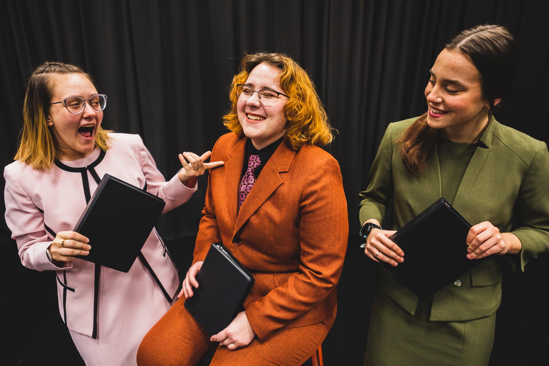 学生Maddy Ramey, wearing a green suit; Olivia Vore, wearing an orange suit with pink tie; and Ali Moulton, 穿着粉红色的西装, laugh in front of a dark curtain while holding small black binders, used for interpretive events in competitive speech.