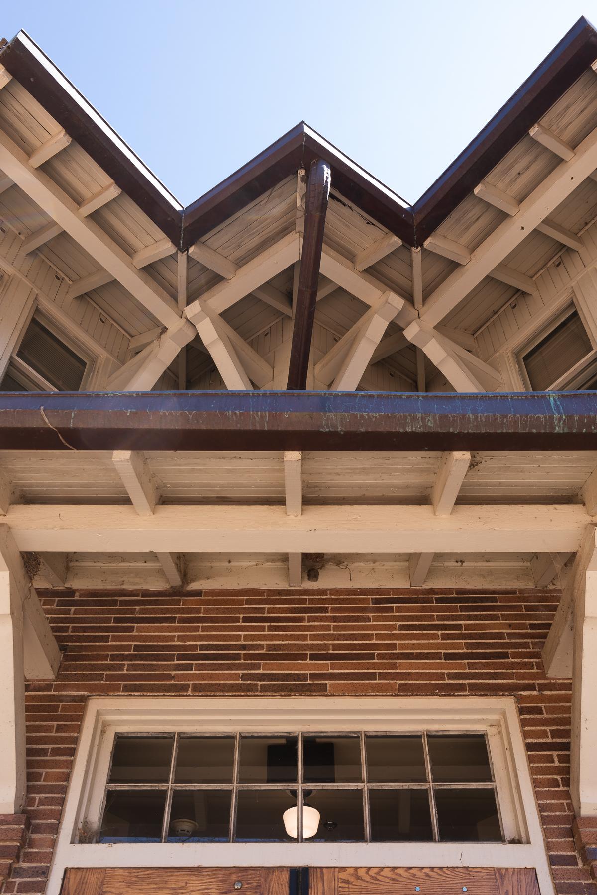 蓝色的天空构成了图像的顶部, dissected into a "W" by the underside of the roof of a building. 在图像中向下移动, there's a metal gutter at the edge of a white-painted overhang topping a brick wall and horizontal window. 在窗户里面，你可以看到一个看起来像老式玻璃灯的东西.