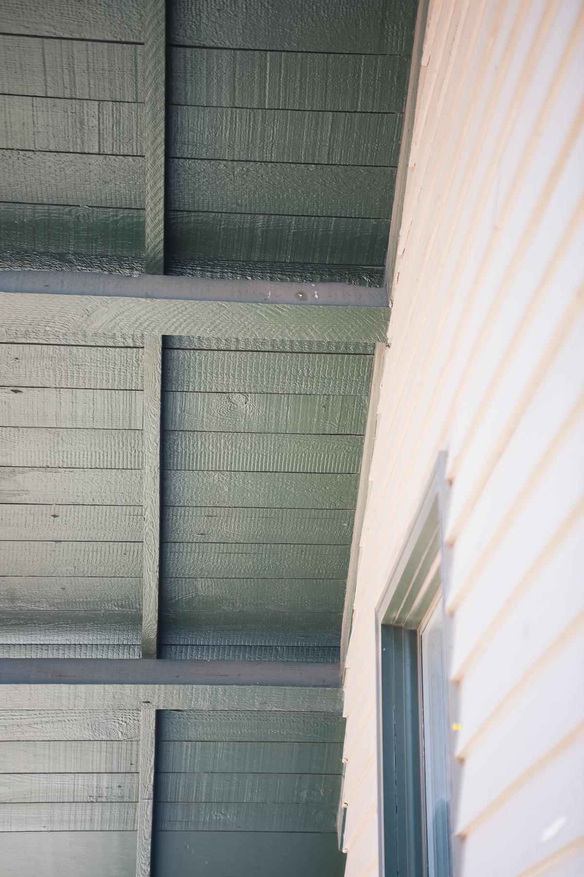 A close-up image of a building shows cream-colored siding and an overhanging, sage green roof. 这座建筑看起来完全是由木头和顶部的双吊顶组成的, 矩形窗口适合照片的左下角.