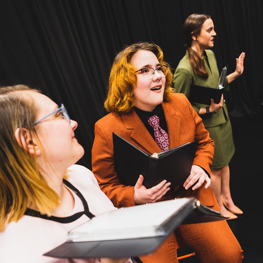 Student Olivia Vore sits on an orange stool in an orange pant suit, 打着粉色领带, holding a small black binder used for interpretive speech events. To their left stands Maddy Ramey in a green skirt suit. On their right, Ali 默尔顿 stands in a pink suit and looks back at Vore. The three students are in front of a dark curtain on a black floor.