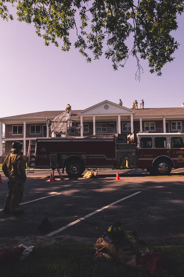 A firefighter watches from the ground as others practice cutting ventilation holes in the roof of Burrage大厅. Jackets and helmets are piled on the ground as other firefighters take a break from the June heat. 