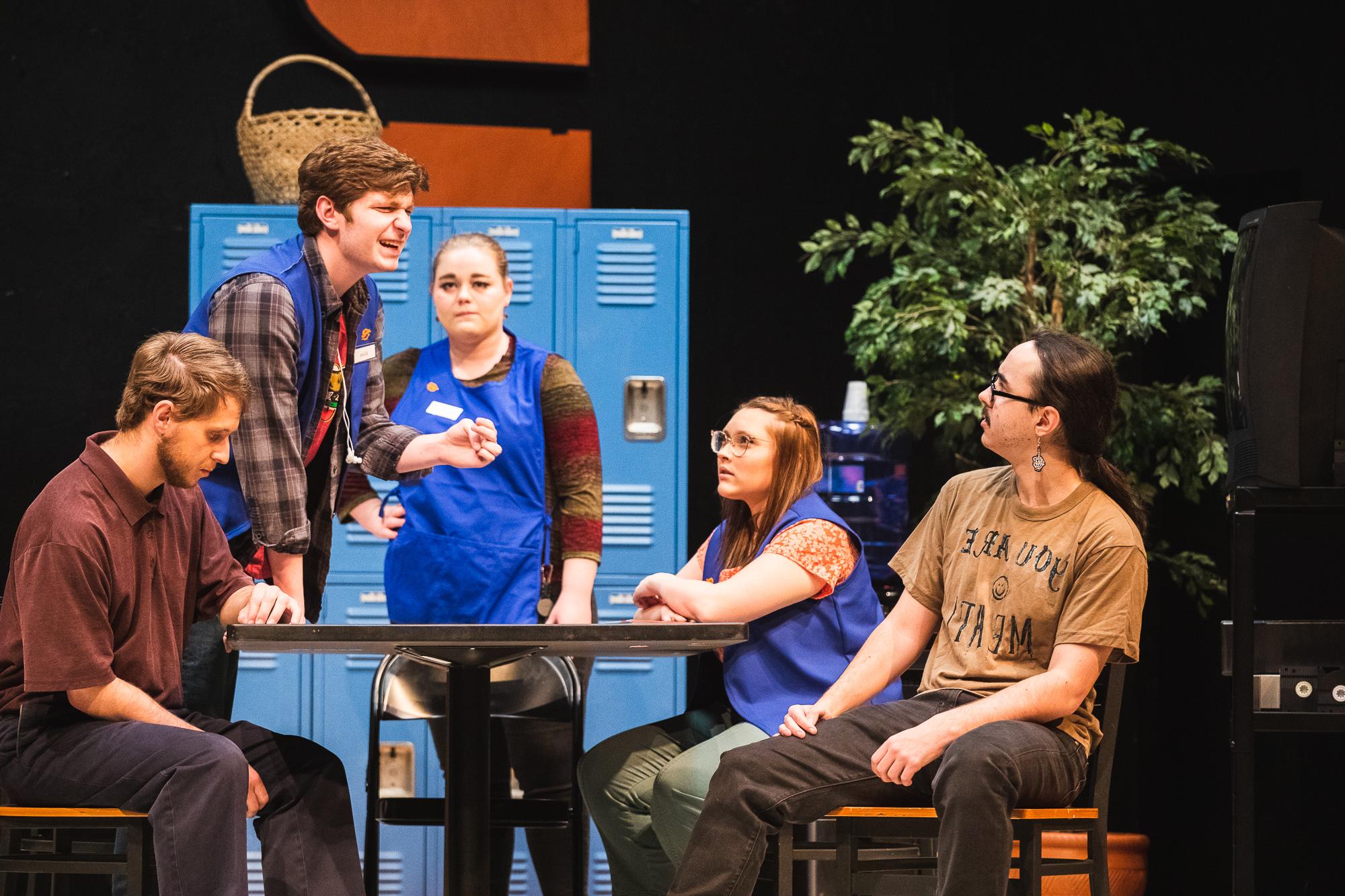 Five actors gather around a table, one standing over the table, clearly distraught. Behind them is a set made to look like the back room of a Hobby Lobby, with a tv on a stand, a sad plastic plant, bright blue employee lockers and a crafty-looking wicker basket. 