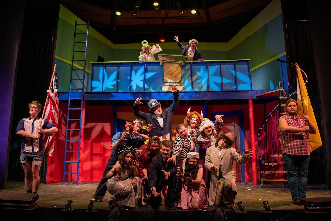 An image from the production of "Reefer Madness" showing actors on a two-level set with brightly colored lighting shaped to look like marijuana leaves. 两个演员, one in a fur cap with a tail and one in an angel costume, stand on the top level of the set. 在基本水平上, two actors stand on the far left and right with flags, and a group of 11 actors pose together in the middle. 