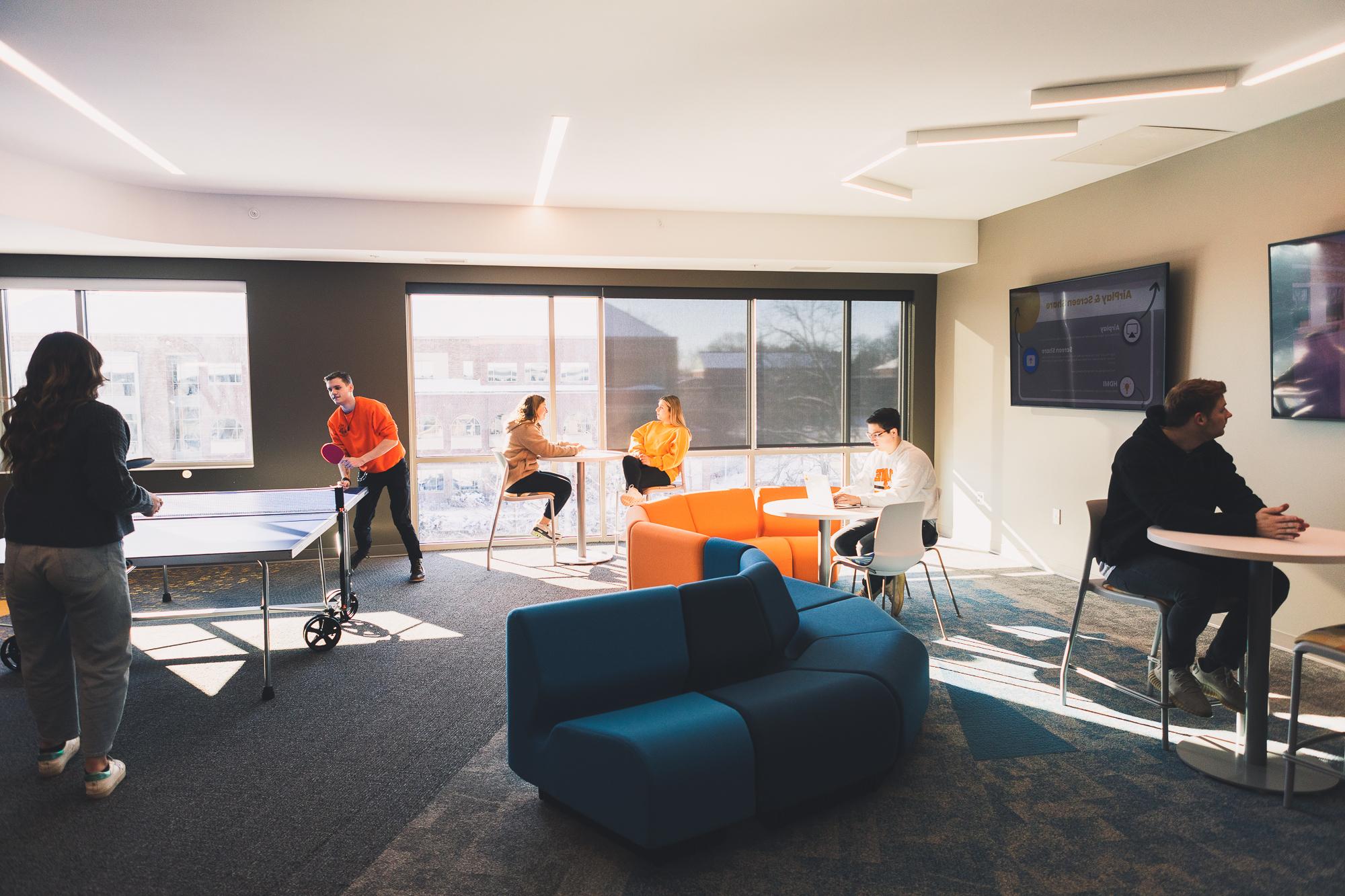 Students use a lounge in New Hall, which is well-lit by a bank of windows. Two students play ping-pong to the right, while two other students sit and talk at a high-top table near the windows. A third student works on a laptop at a small, white, round table around which an orange couch curves. 