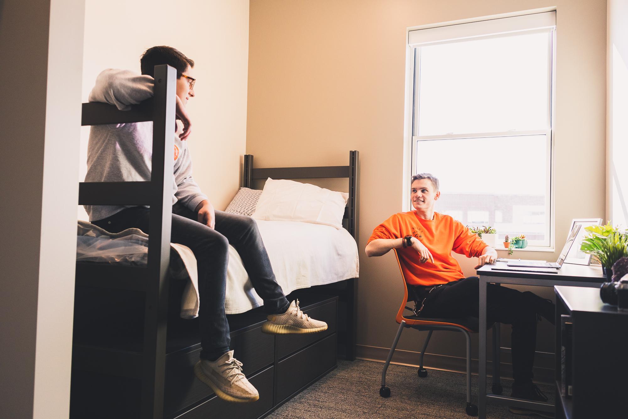 Kaidan Smith looks up from a desk at Trenton Johansen on the bed of one of the third-floor suites. Also in the room is a small bookshelf and the window looks out onto Doane's campus. 