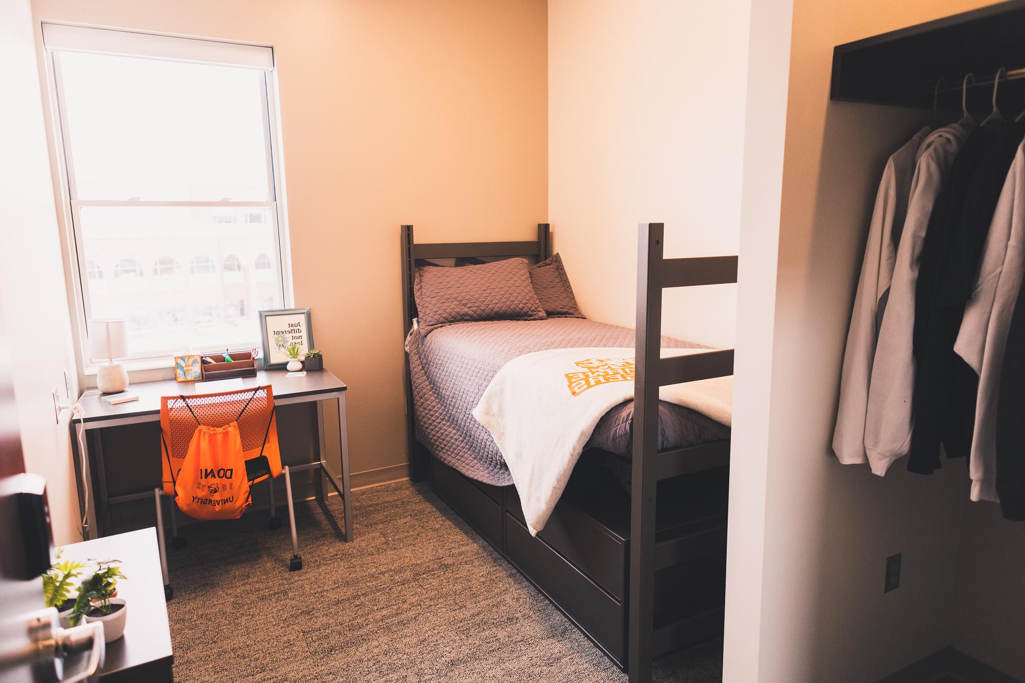 A sample room decorated with a Doane Tigers blanket and other Tiger promotion items. A desk is pushed up against a window, and a bed along the wall to the left of the desk. 