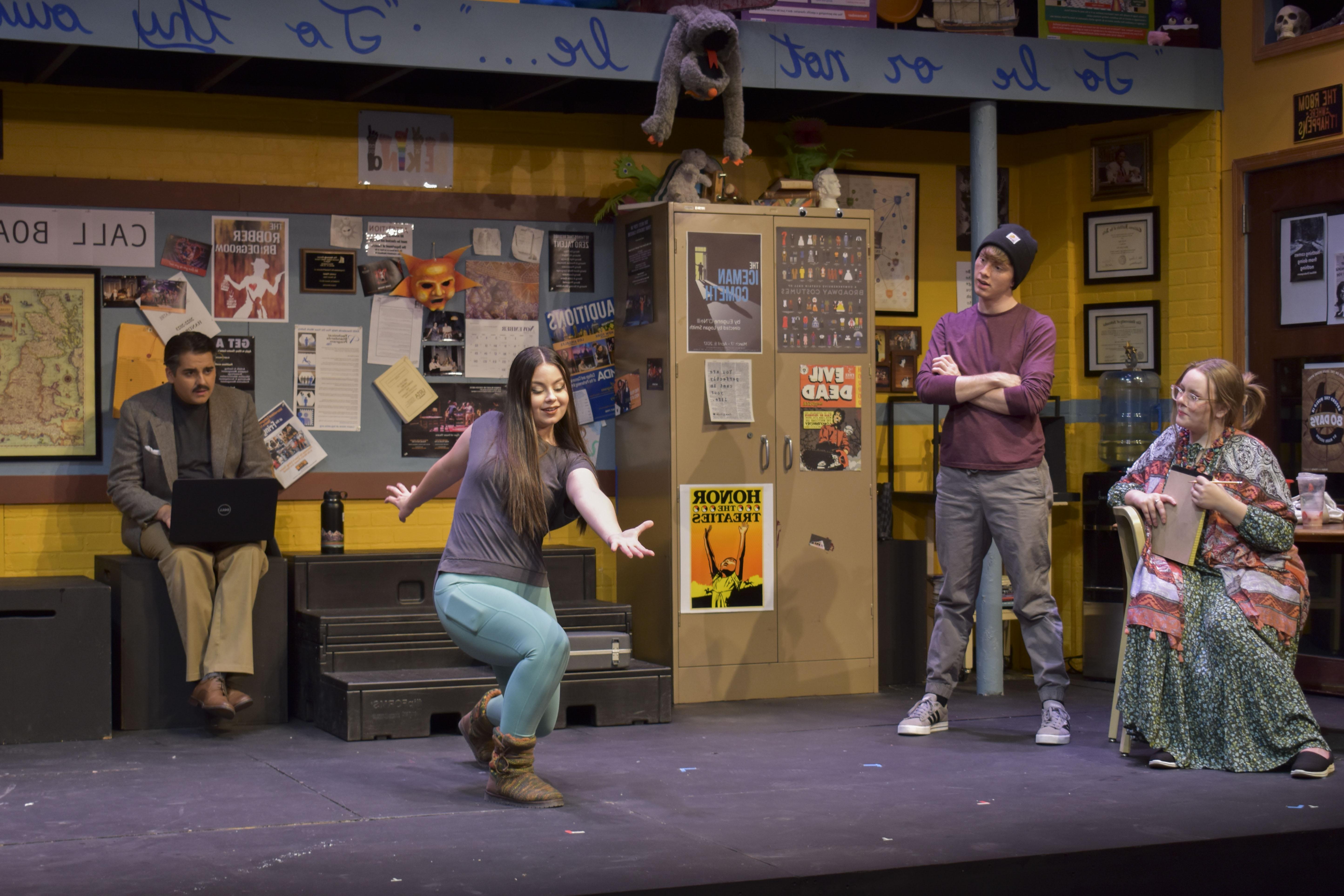 Four actors pose on a stage, with one miming a bowling motion. The stage has two levels, and a puppet dangles precariously from the second above the words "To Be or Not To Be." The rest of the set looks like a 剧院 classroom, 有桌子和椅子, posters covering the walls and props littered around the edges.