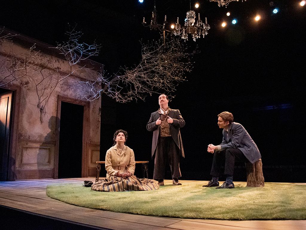 Image of a scene from 澳门威尼斯人网址剧院's spring 2023 production of "樱桃园." Three actors sit or stand on a rug of artificial turf on the set of the production, which combined natural elements like tree branches with walls, furniture and chandeliers that show hard times falling on a bourgeousie family.