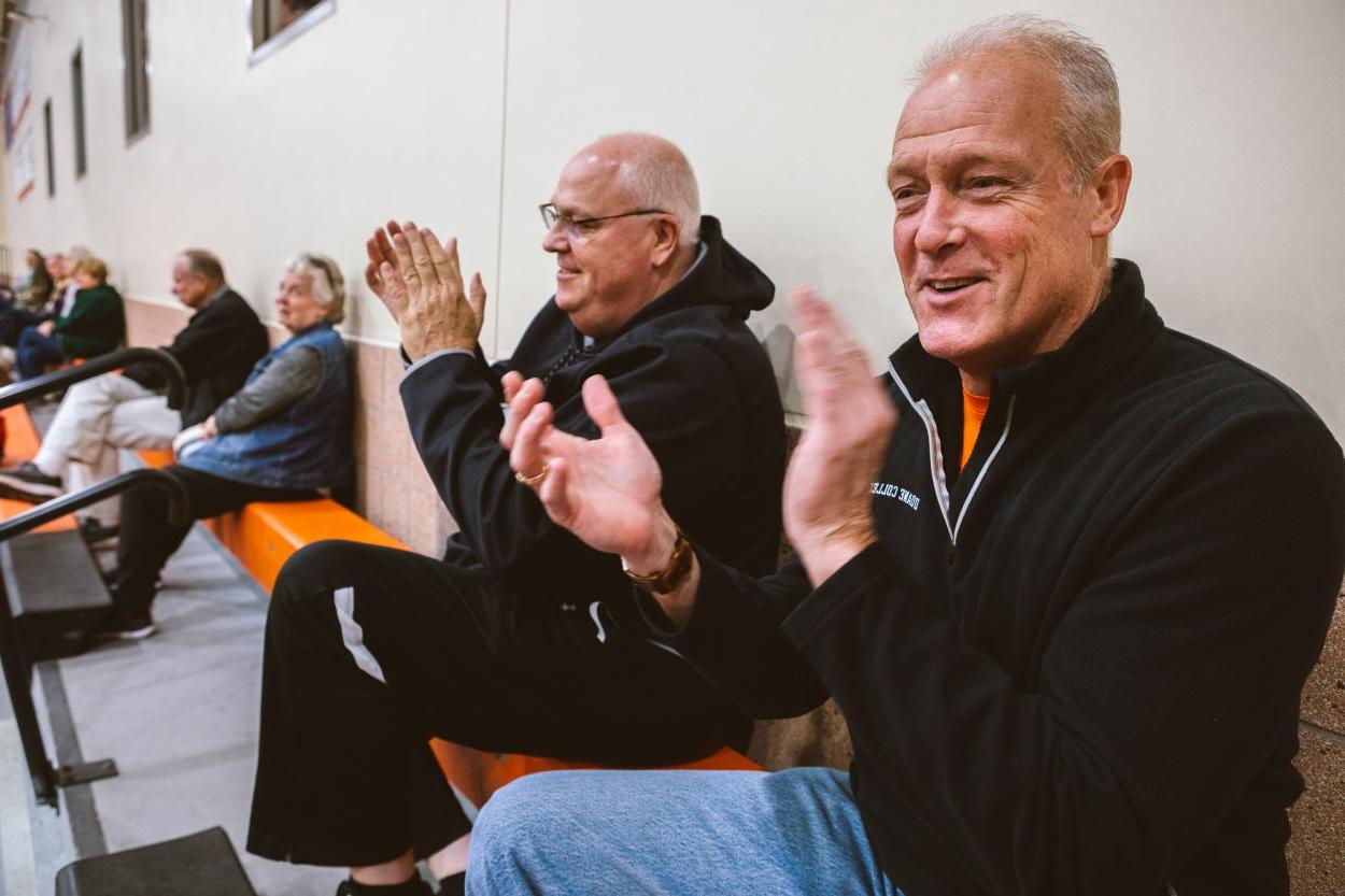 Tom Sorensen (left) claps for the Doane Women's Basketball team. He wears a black quarterzip with a "Doane College" logo, and sits at the top of the orange bleachers next to Marty Fye.