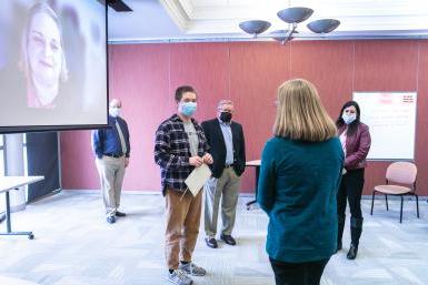 Samuel Province (middle, plaid shirt) speaks with Nancy Murphy, financial aid counselor. He holds a certificate announcing his scholarship. The donor for whom the scholarship is named, Carolyn Kollmeier '74, joined by Zoom and is shown on a screen on the right side of the image.