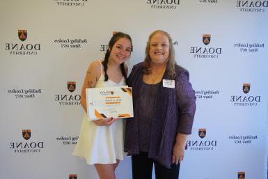 A woman in a purple shirt and jacket stands next to a woman in a short white dress holding up an award printed on white paper. The two stand in front of a white backdrop with text. 